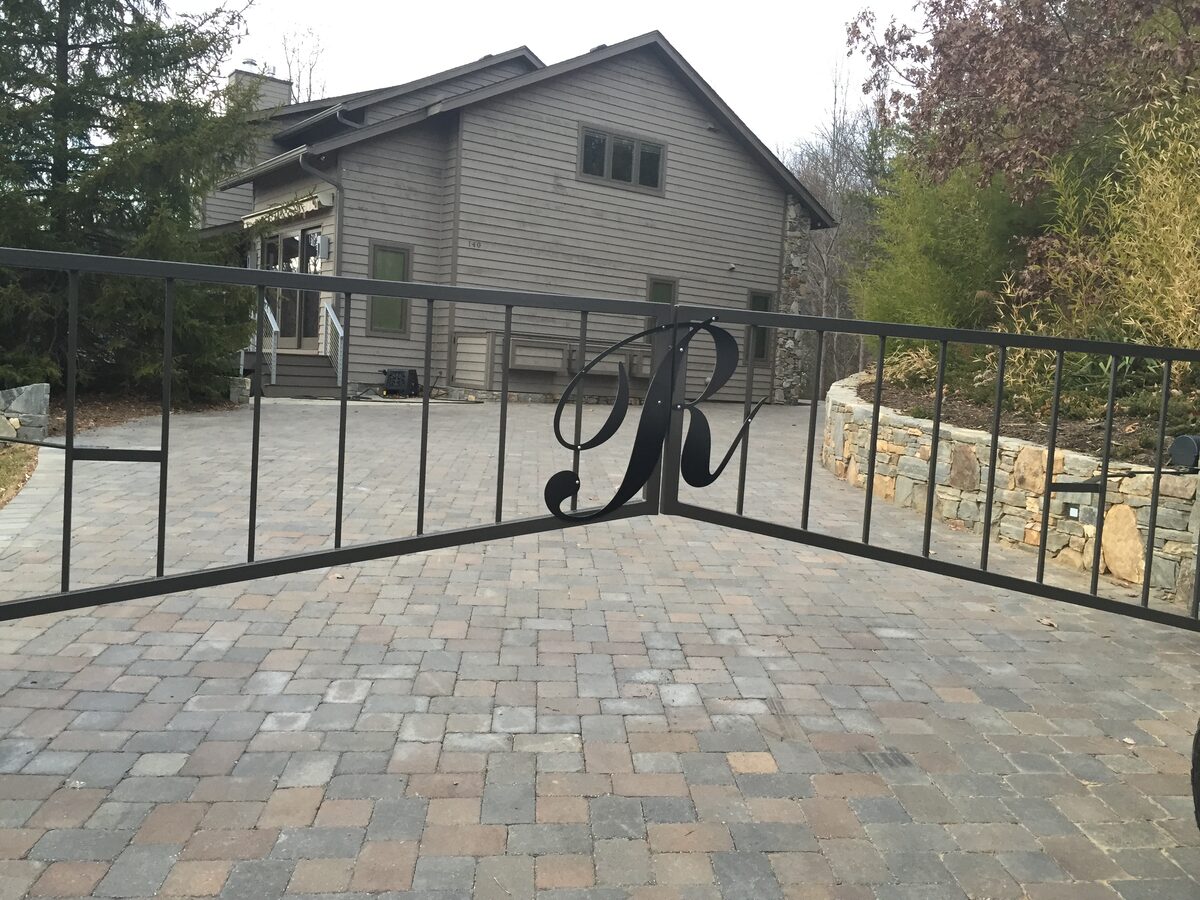 Black ornamental fence section with a custom fabricated script monogram over a paver drive