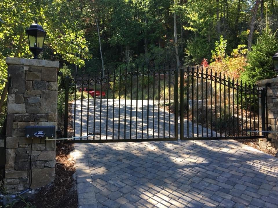 Swing gate operator mounted on a stacked stone pillar with the drive arm connected to a black ornamental double gate over a paver driveway
