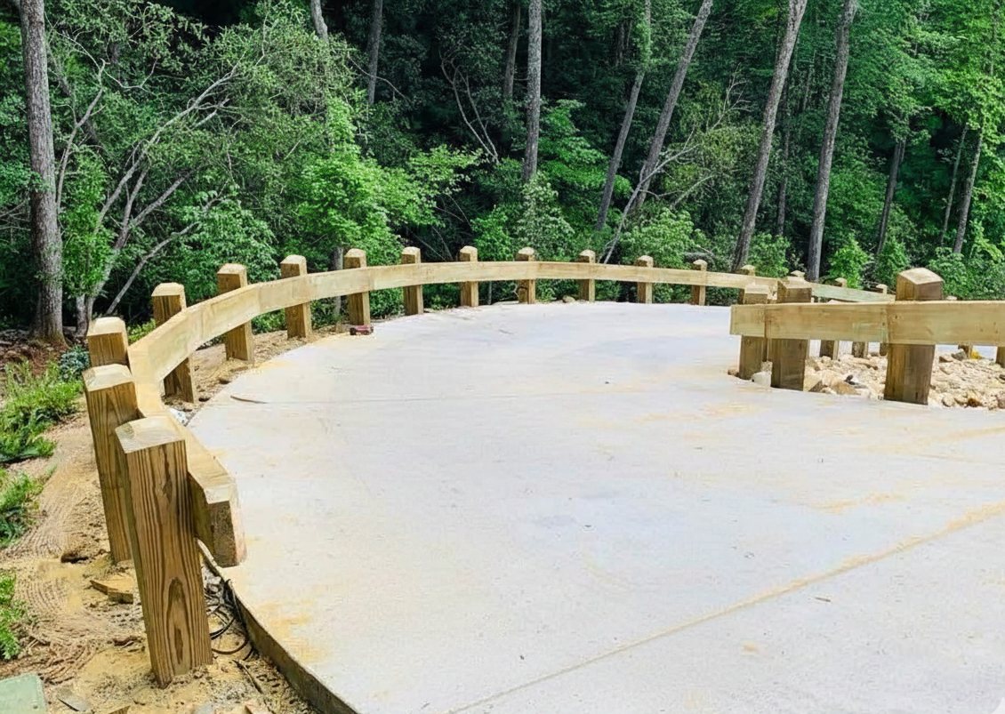 Curved timber post-and-rail guardrail set along the edge of a new concrete parking pad in the woods