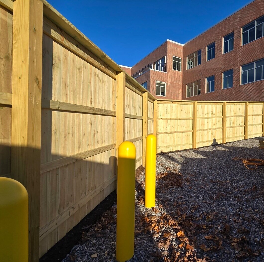 Tall pressure-treated wood privacy screen with yellow bollards alongside a brick commercial building